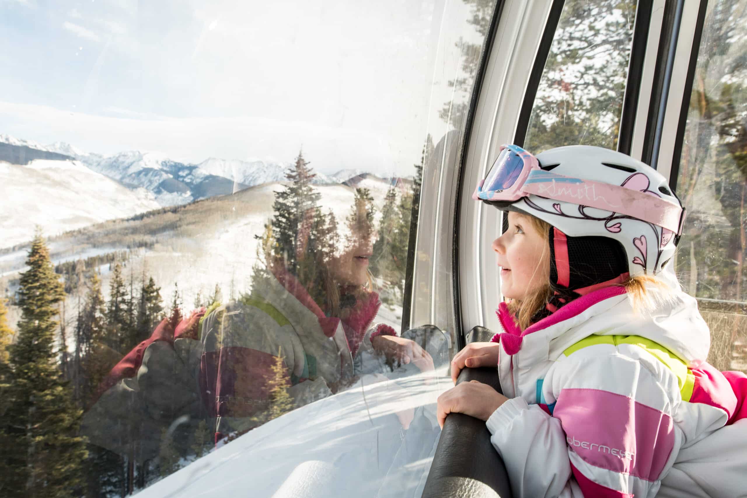A child wearing a white and pink ski helmet, googles and jacket stares at Vail winter scenery from the gondola window.