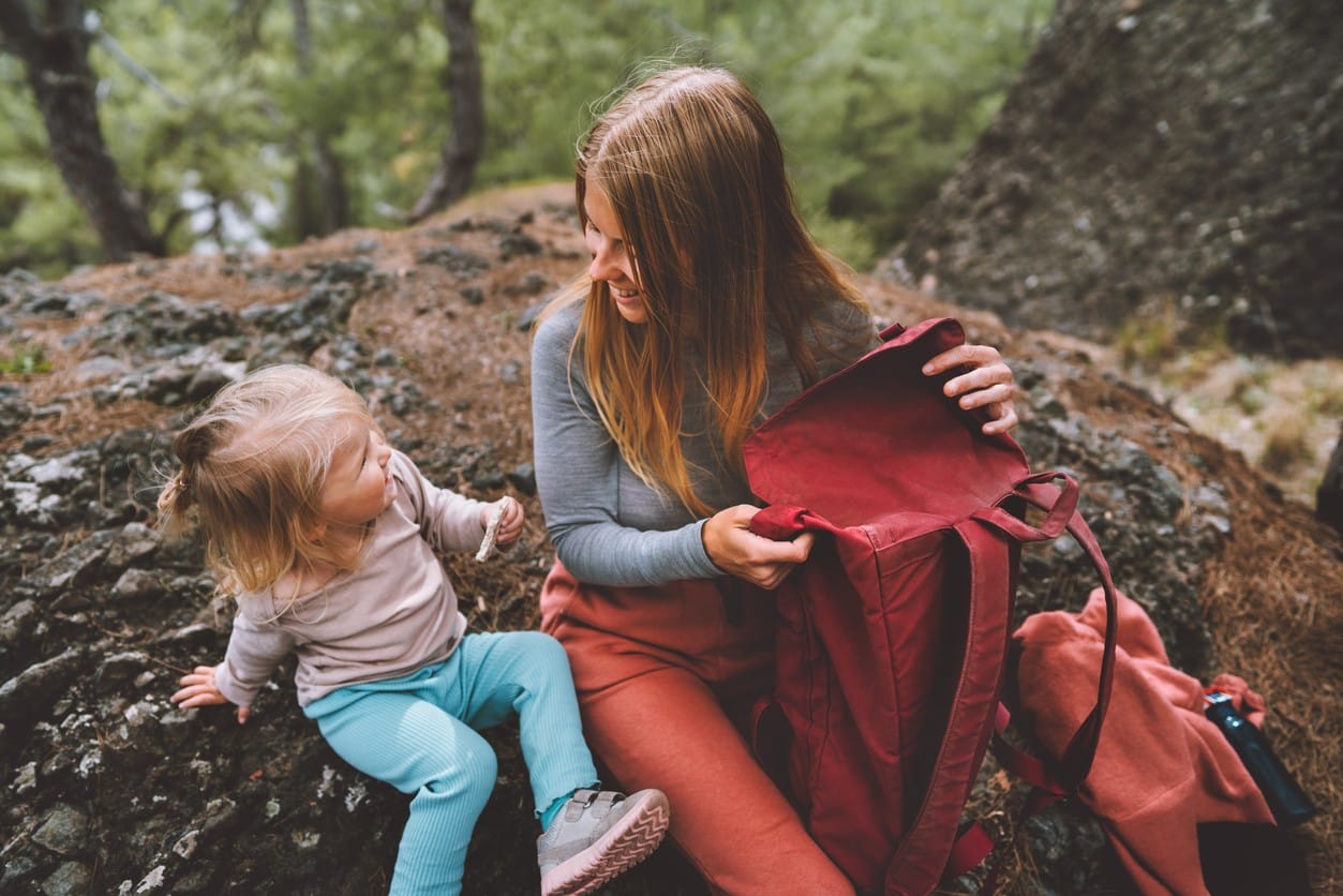 A mother looks at her child smiling as they sit on a rock along a trail in Vail with a red backpack half open.