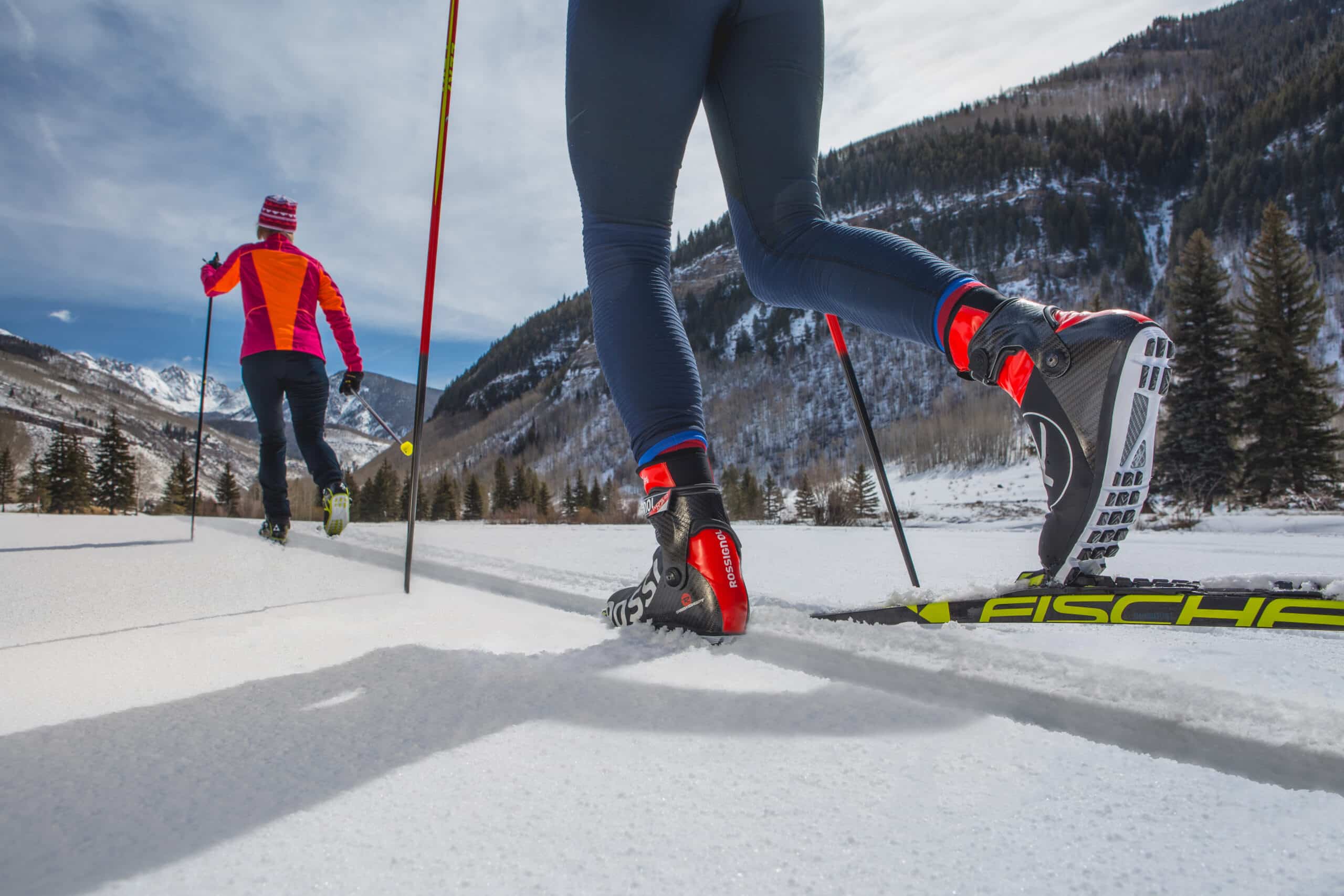 Two people cross-country ski through a Vail valley. The person in front wears an orange and maroon winter jacket.