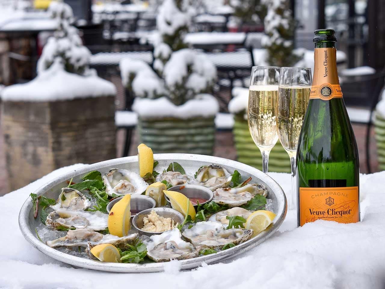 Oysters on a platter with two glasses of champagne and a champagne bottle in snow outside Vail Chophouse.